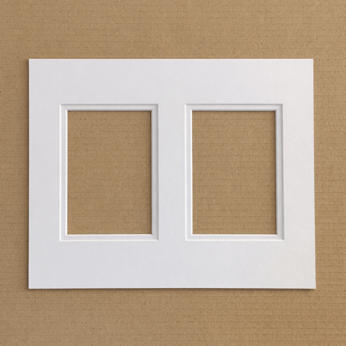 A white double-frame mat board or picture frame mat with two rectangular recessed openings, shown flat against a brown textured background.