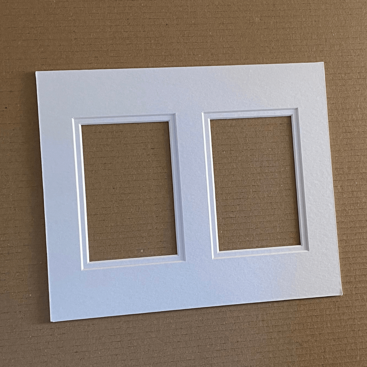 A white double mount board with two rectangular inner cutouts, photographed flat on a brown corrugated cardboard background.
