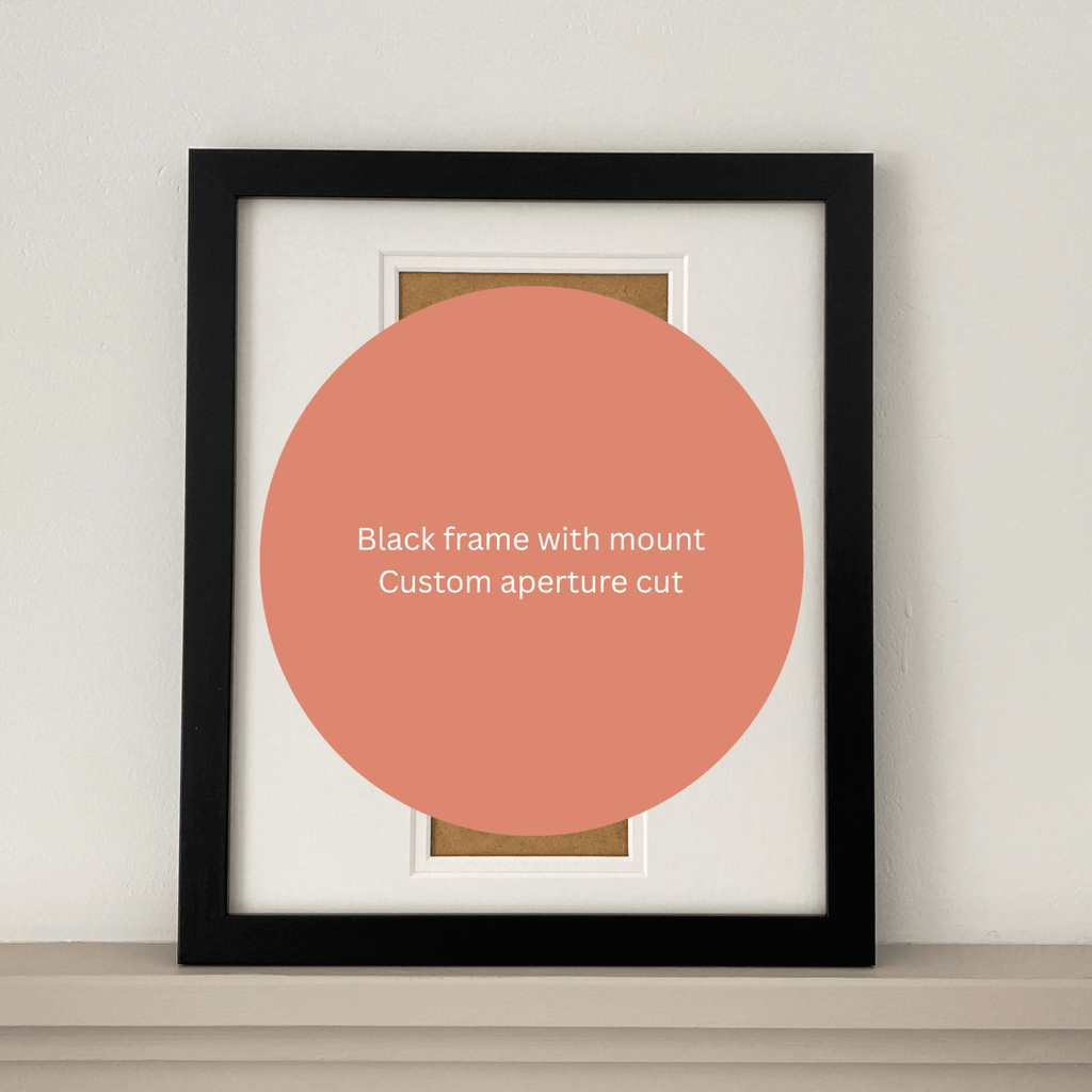 A black picture frame with white matting and a tan backing board, leaning against a light wall on a shelf. A large peach circle overlay in the center reads, 'Black frame with mount Custom aperture cut.