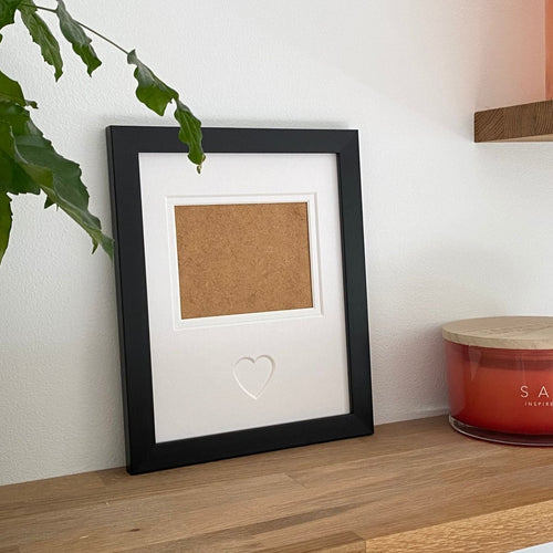 Black single-aperture heart picture frame displaying family photo on wooden shelf