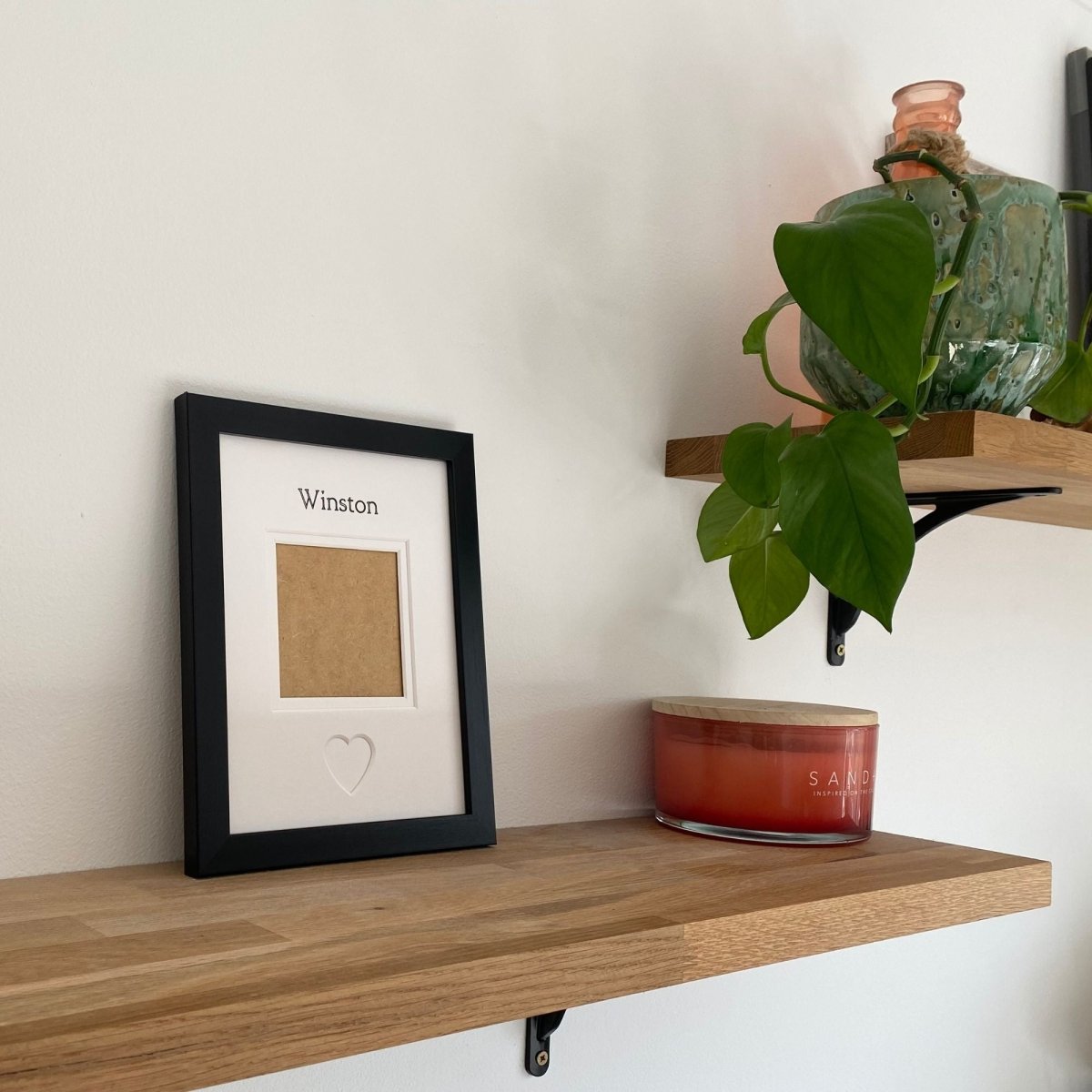 Wooden shelf with a framed picture, candle, and plant against a white wall.