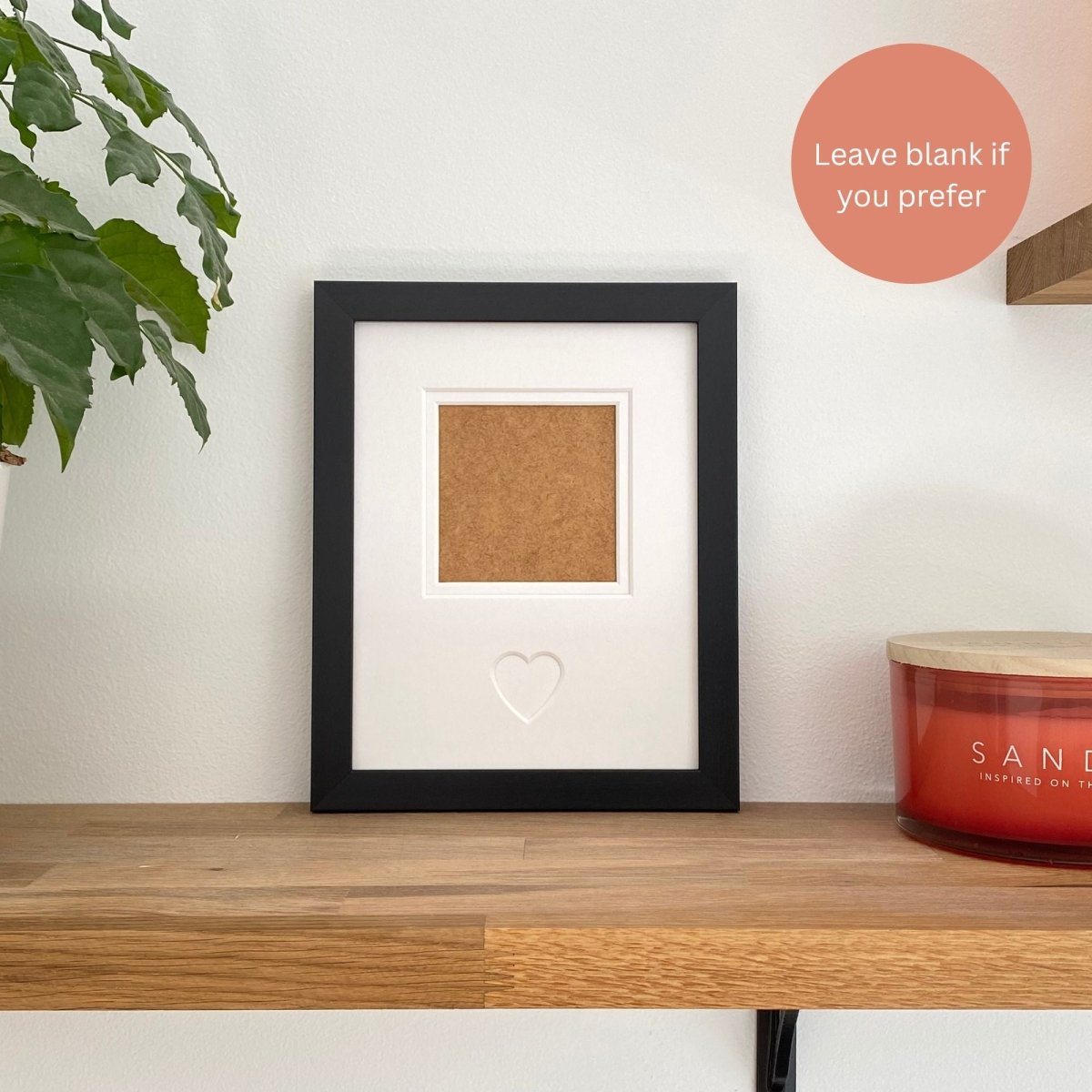 Framed with a single aperture with a heart cutout on a wooden shelf next to a plant and candle.