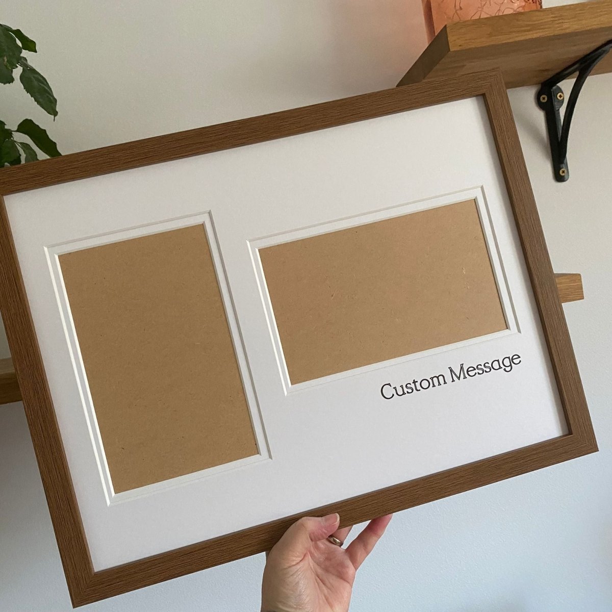 Hand holding a large brown frame with white double matted, two aperture cut-out by a wooden shelf and white wall