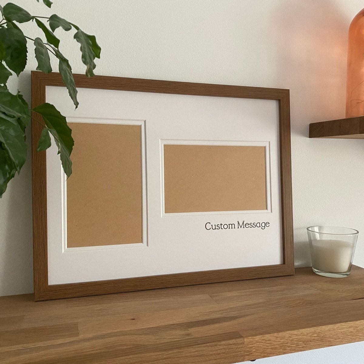 Large brown picture frame standing on the wooden shelf next to a white candle and green leafy plant againt a white wall and rosy pink glass vase