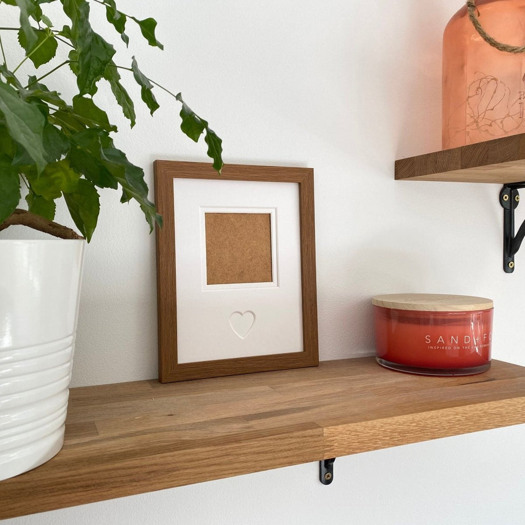 Brown picture frame displayed on a brown shelf beside a red candle, a white plant pot, and a rose‑coloured vase