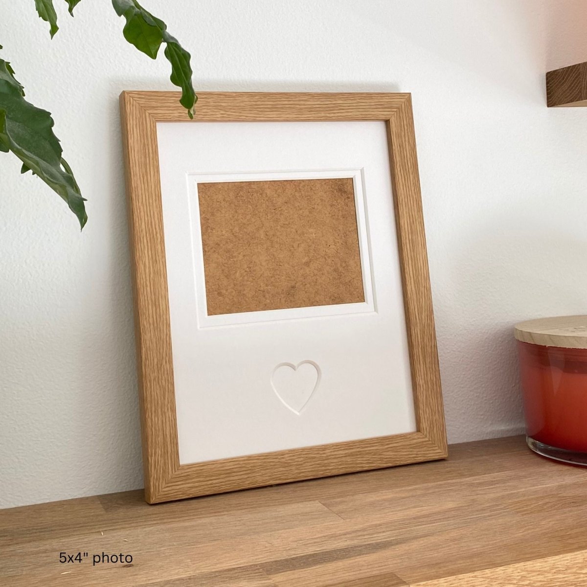 Light brown photo frame with a heart cutout on a wooden surface with a plant and candle in the background.