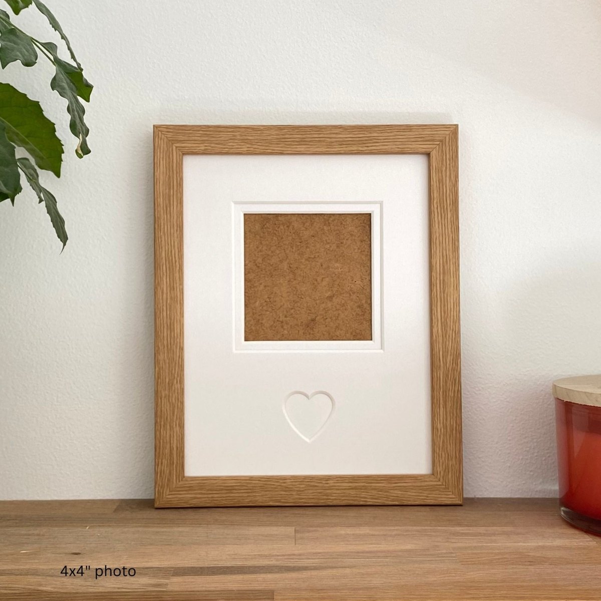 Light brown photo frame with a heart cutout on a wooden surface with a plant and candle in the background.