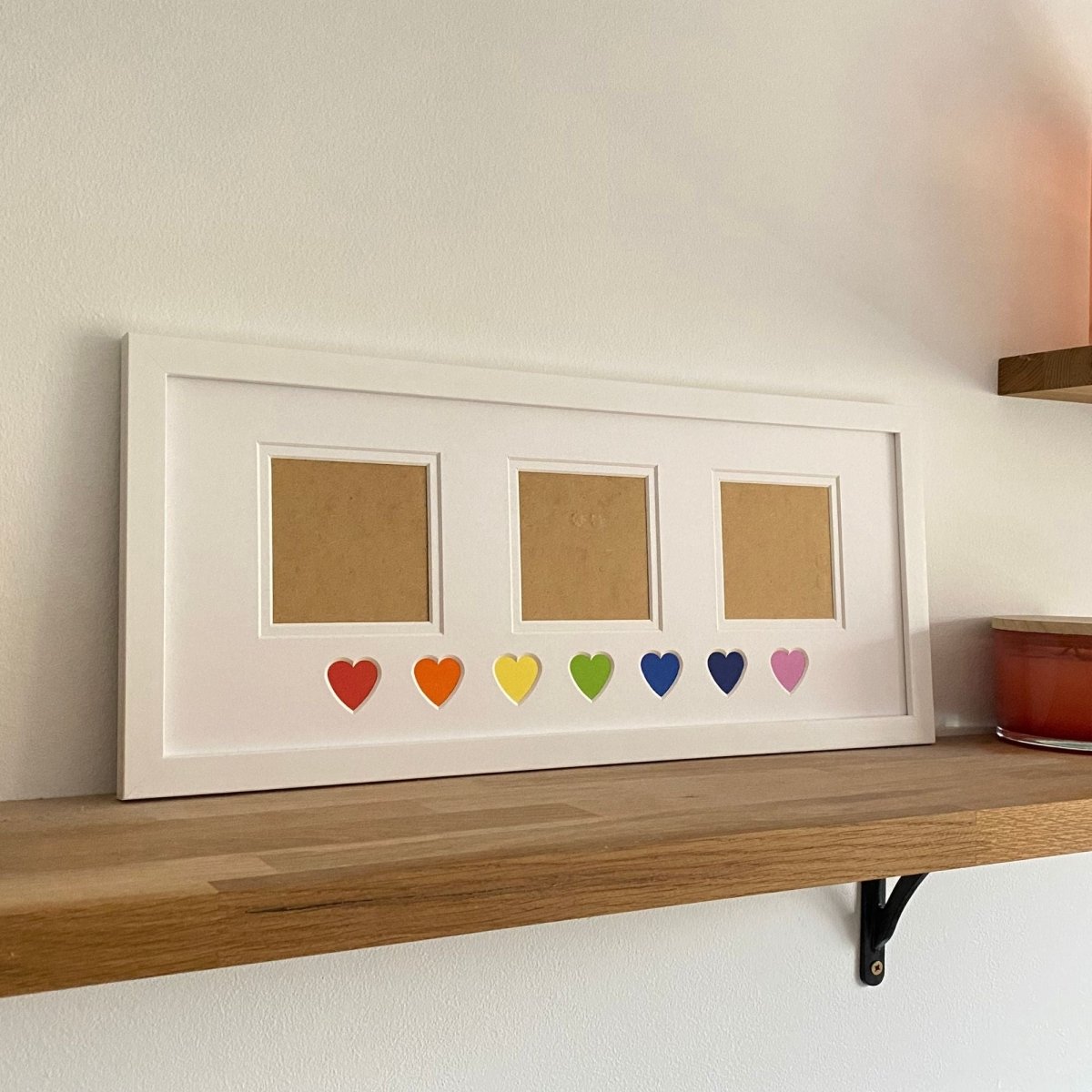 White photo frame with three slots and colourful hearts on a wooden shelf.