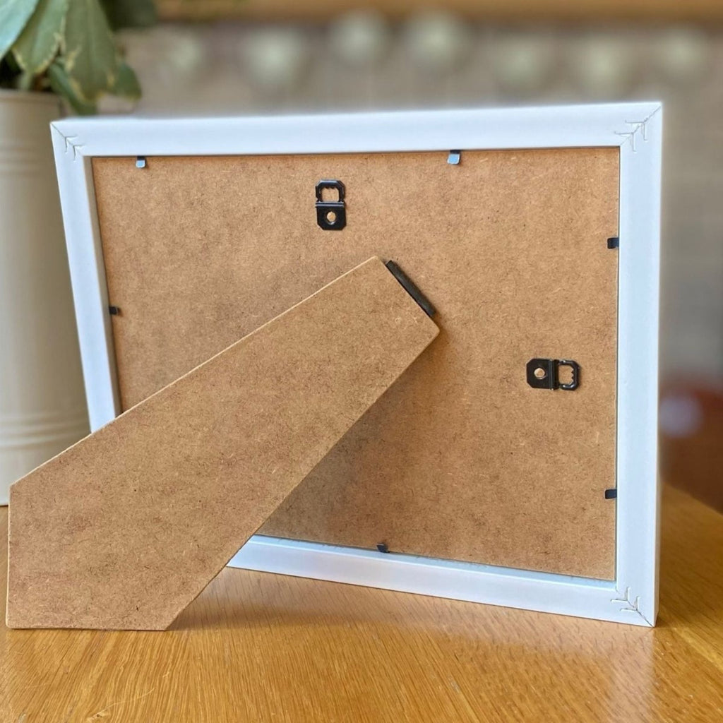 Back of a white picture frame with wooden backer on a wooden surface.