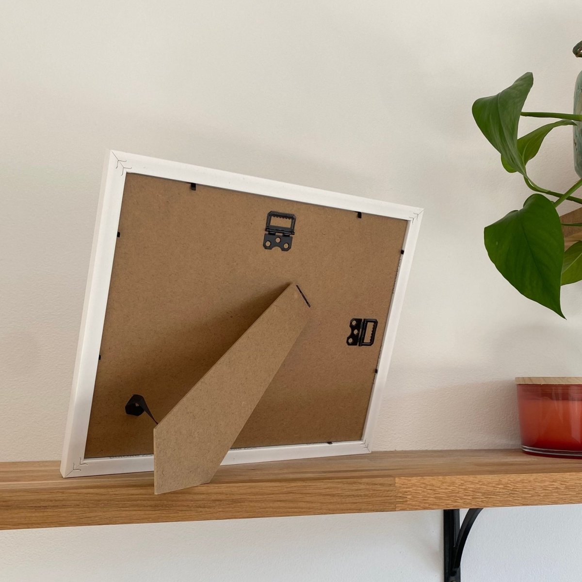 Back of a white frame on wooden shelf, light wall, green leaf and red candle 