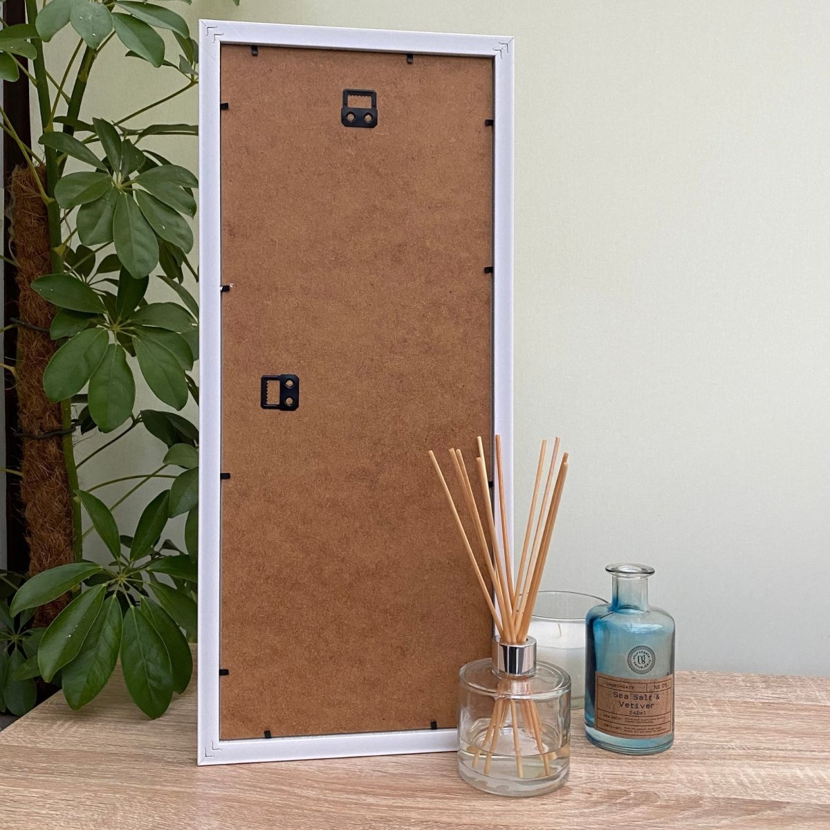 Back of white frame on the tabletop next to a diffuser, candle and green plant