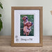 Freestanding, light brown contemporary picture frame on a tabletop with a plant. An image of the fox red puppies, siblings, brother and sister on the grass.