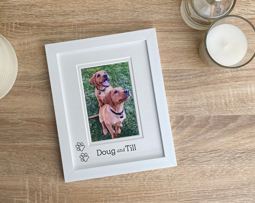 white photo frame laying on the tabletop next to a white candle and diffuser. The image inside the frame is two fox red labrador puppies.