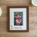 dark brown picture frame laying on the tabletop next to a candle, jug and a diffuser. An image inside the frame is a fox red labrador siblings with their names inscribed on the double white matted.