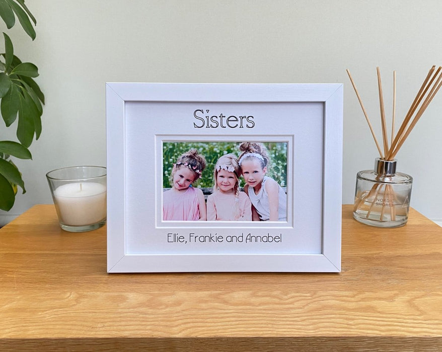 White picture frame, double white matted with Sisters and personalised with all the sister's names. Standing next to a white candle and diffuser on the table top with a plant on the side.