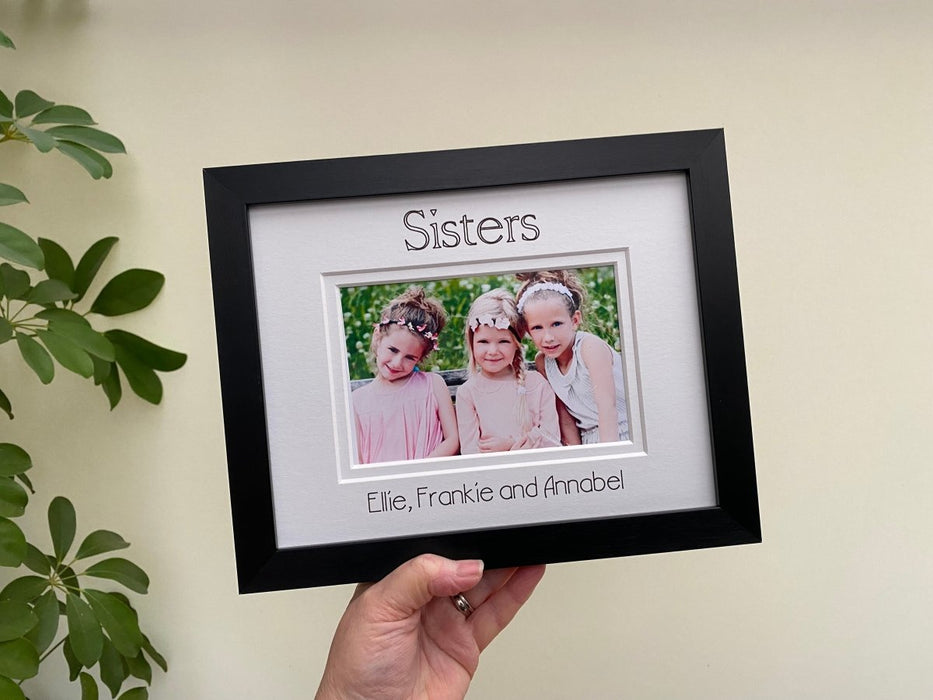 Holding up a black photo frame of the sisters with their names inscribed at the bottom of the white mat. Next to a green plant.