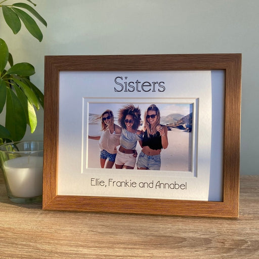 Dark brown picture frame, double white matted with Sisters inscribed and personalised with all the sister's names. Standing next to a white candle on the table top with a plant on the side.
