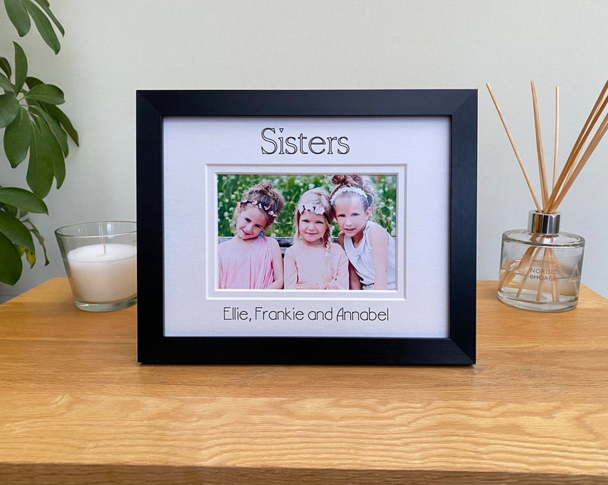 Black picture frame, double white matted with the Sisters inscribed and personalised with all the sister's names. Standing next to a white candle on the table top with a plant on the side.