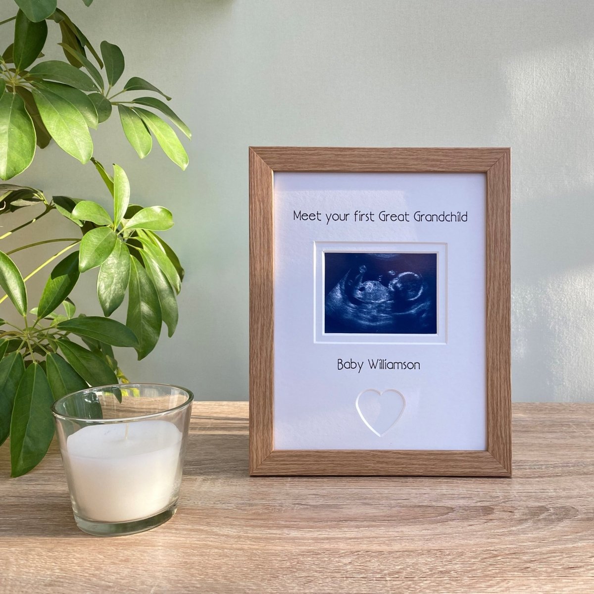 Freestanding, light brown, wood-grain effect picture frame, sonogram scan image of 1st Great Grandchild with a heart shape. Next to a white candle and green plant