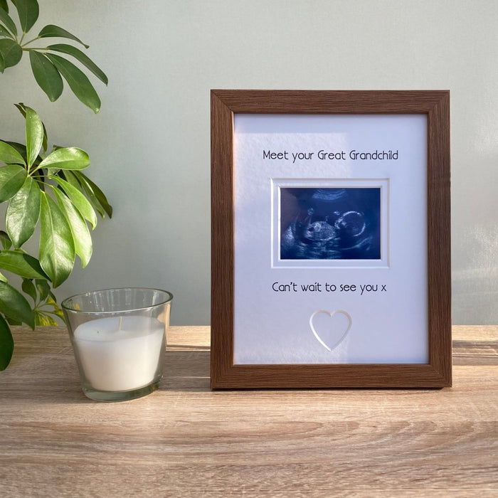 Freestanding, dark brown picture frame, scan image of Great Grandchild with a heart shape. Next to a white candle and green plant