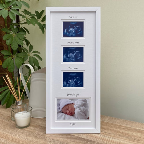 White upright photo frame displaying a newborn baby and three ultrasound scans on a brown tabletop beside a white candle, a cream jug, and a small green plant.